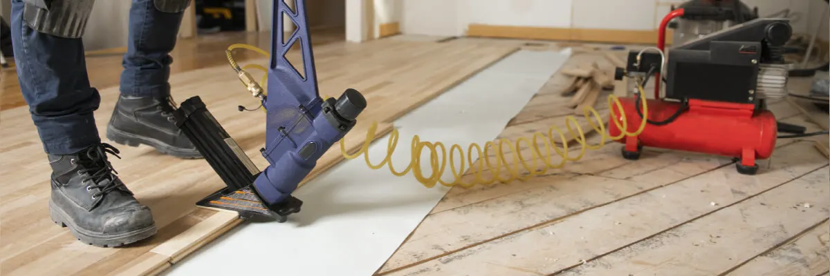 Close-up of a worker's boots using a pneumatic nail gun connected to a red air compressor while installing wooden flooring.