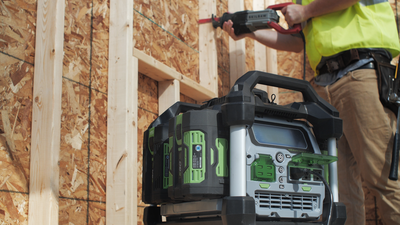 Green and black portable power station with dual 7.5Ah batteries powering tools on wood frame construction site