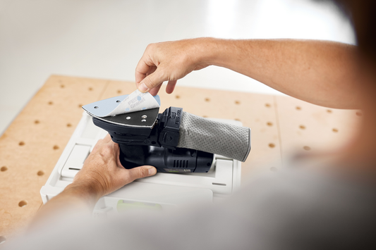 Person attaching Festool Granat Delta abrasive sheet to compact power sander in home improvement workspace