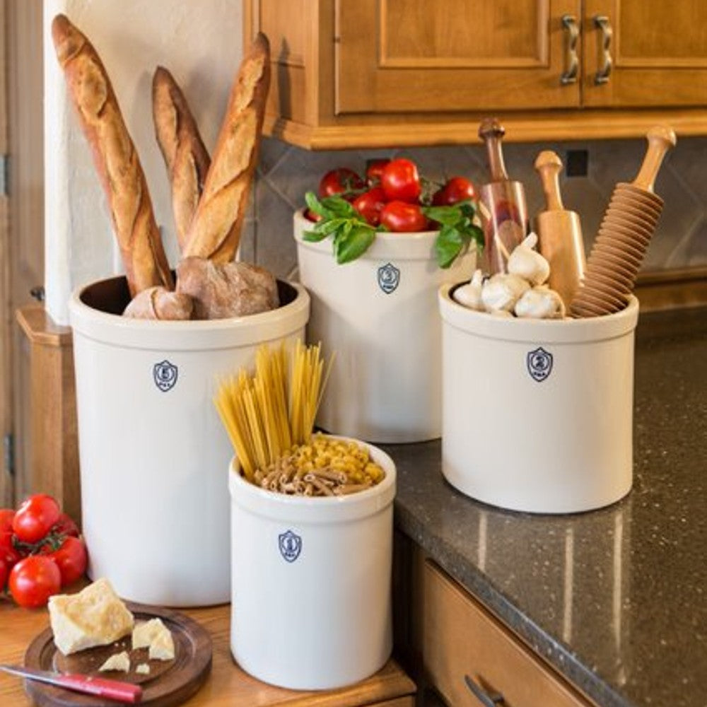 Set of beige stoneware crocks with blue shield emblems holding bread, tomatoes, pasta, garlic, and kitchen tools on countertop