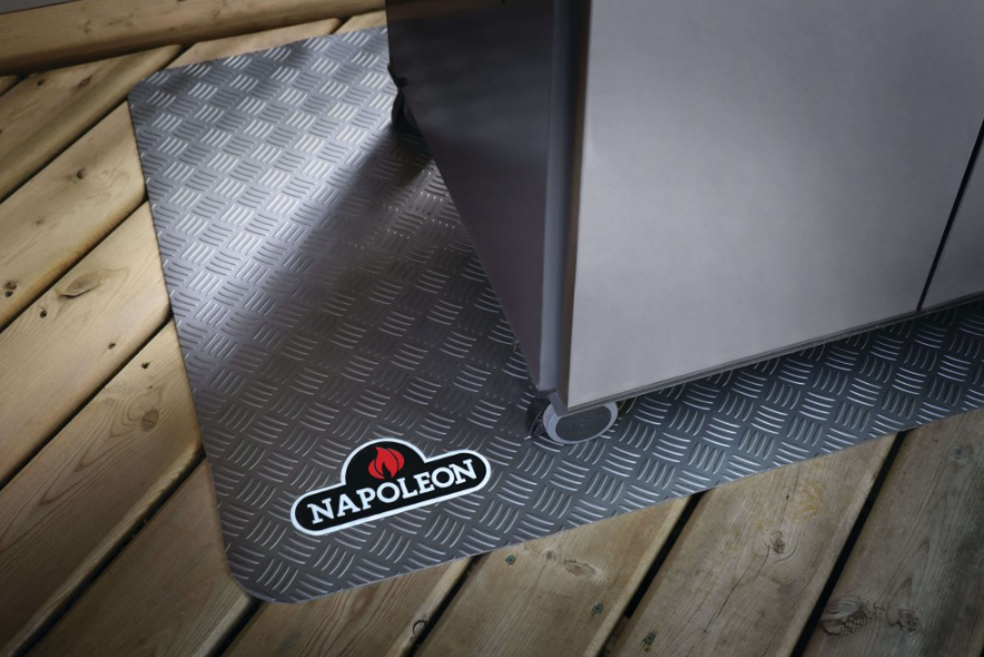 Dark gray grill mat with subtle checker plate texture and rounded edges placed under a stainless steel barbecue grill on wood decking, protecting the surface and showcasing a small branded logo, ideal for outdoor living and grilling accessories.