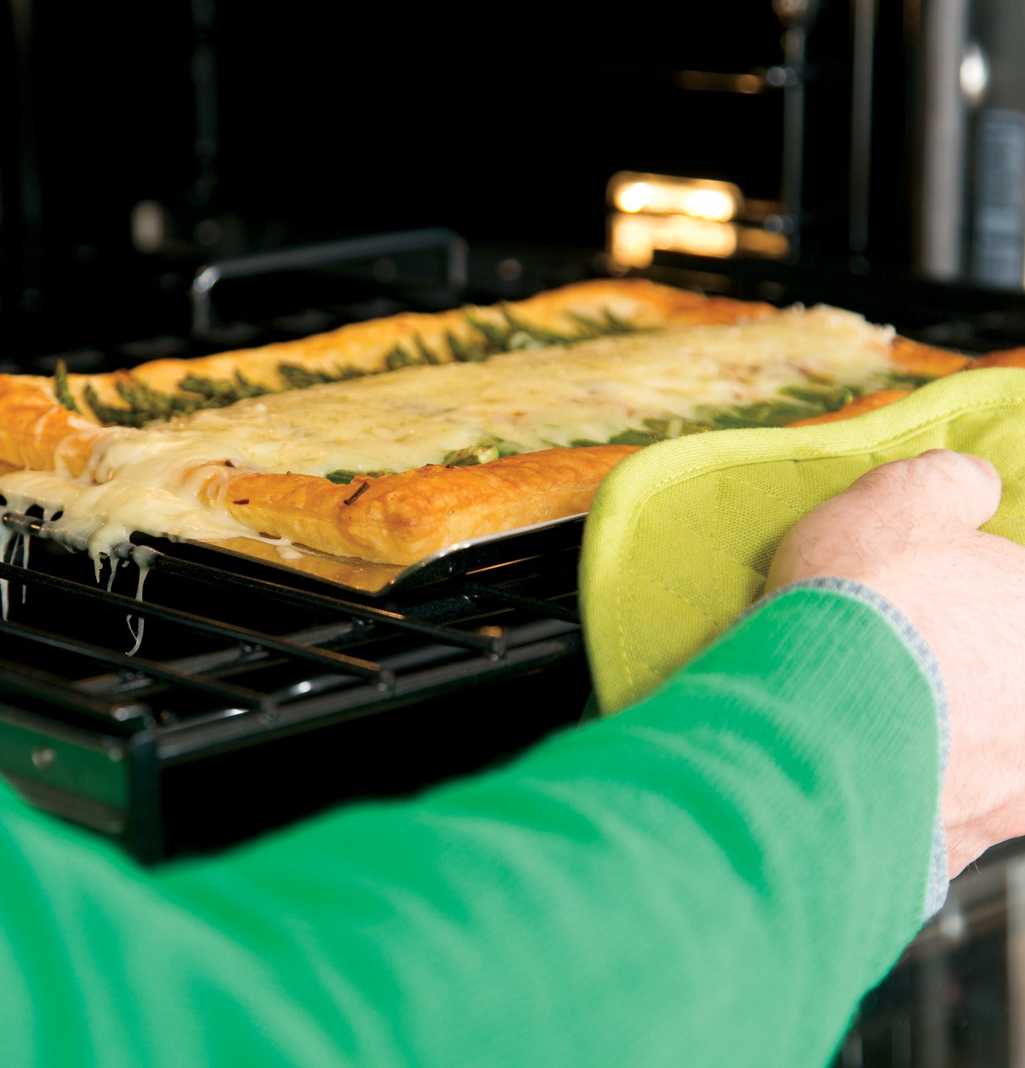 Person wearing green sleeve removing cheesy baked dish from convection wall oven in kitchen and home improvement setting