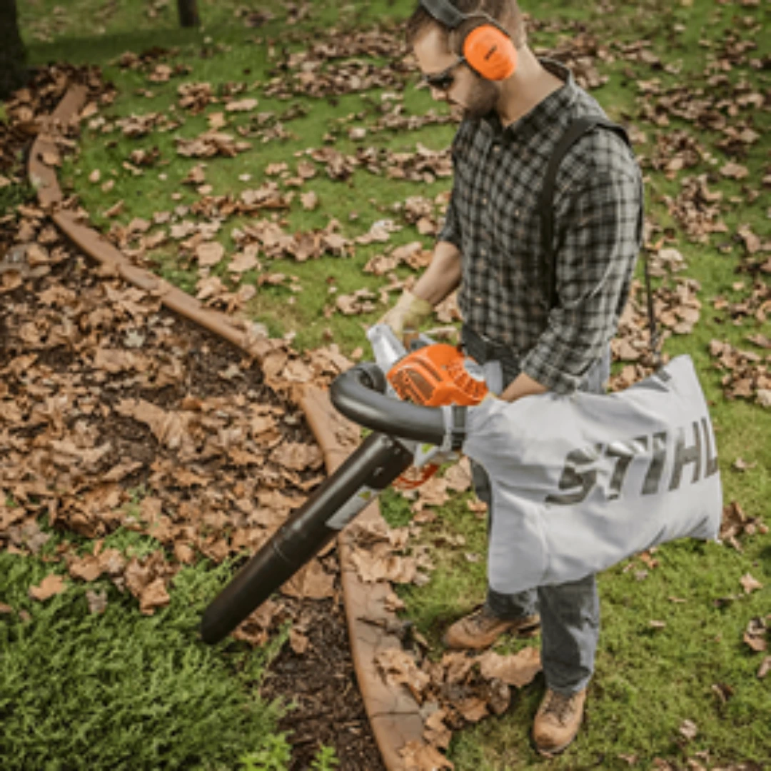 Man wearing protective gear using an orange and white gas-powered leaf blower to clear fallen leaves outdoors