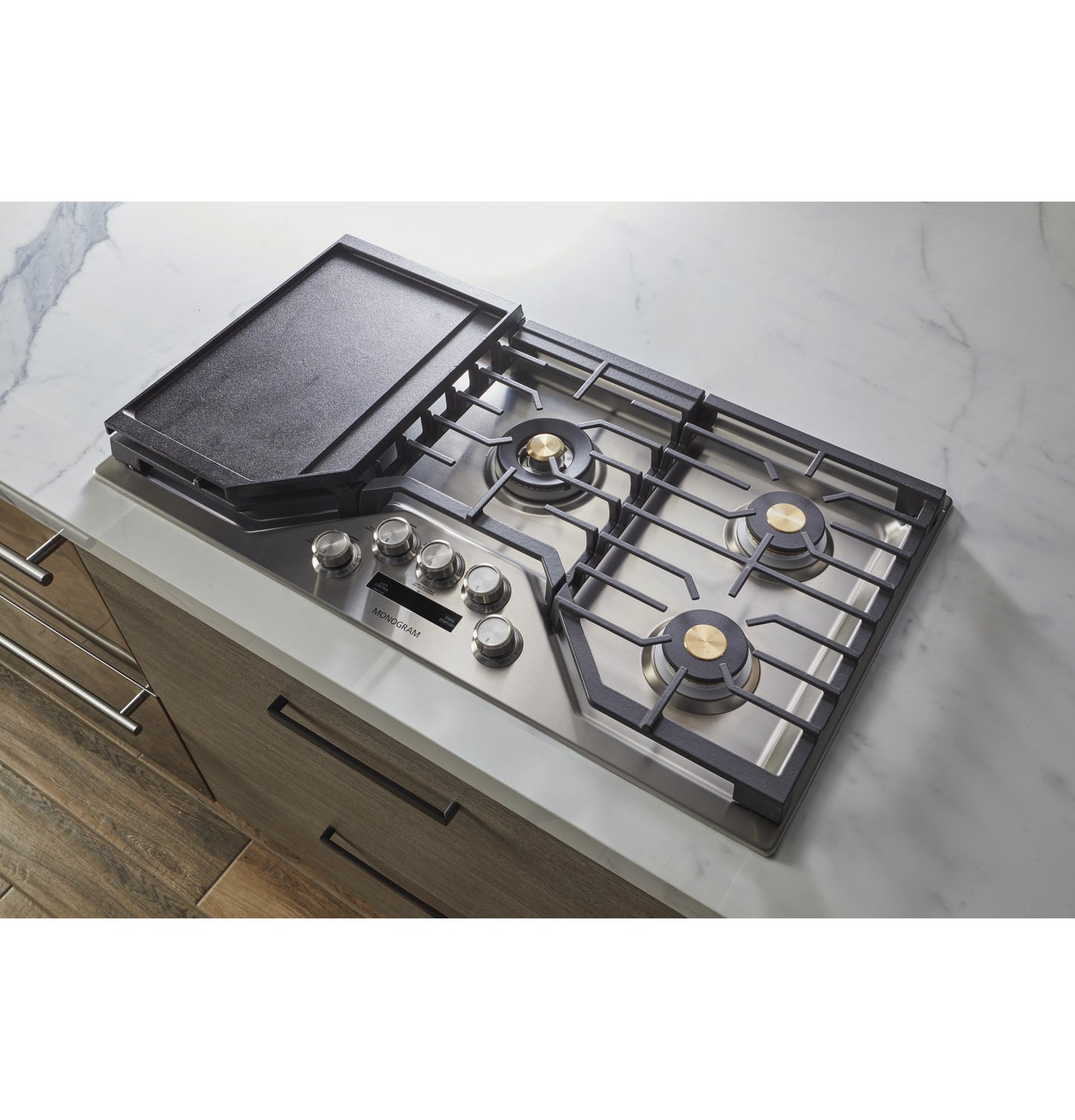 Stainless steel recessed gas cooktop with brass burners, cast iron grates, control knobs, and a cast iron griddle on marble countertop