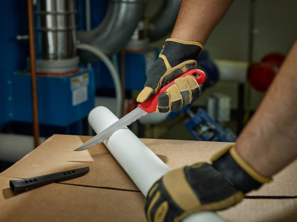 Hand wearing gloves using a serrated blade knife with red handle to cut insulation roll in a workshop setting