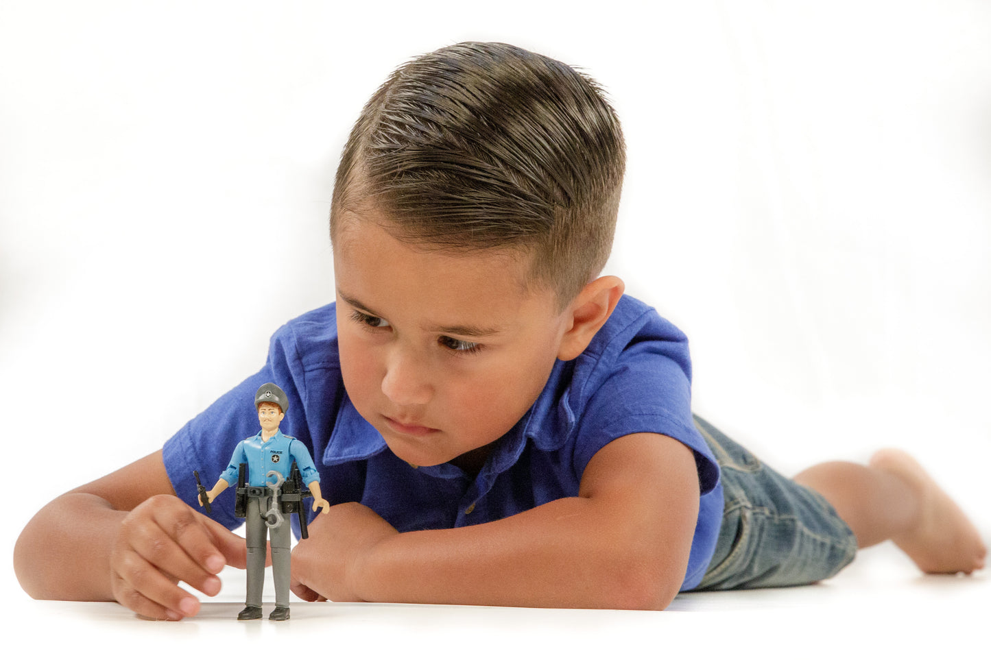 Young boy in blue shirt lying down playing with light skin police figure with baton and belt accessories