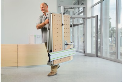 Man carrying folded Festool multifunction table in workshop area with large windows and wooden panels nearby