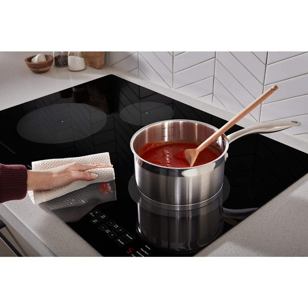 Black induction cooktop with four cooking zones, simmering tomato sauce in stainless steel pot and hand wiping surface