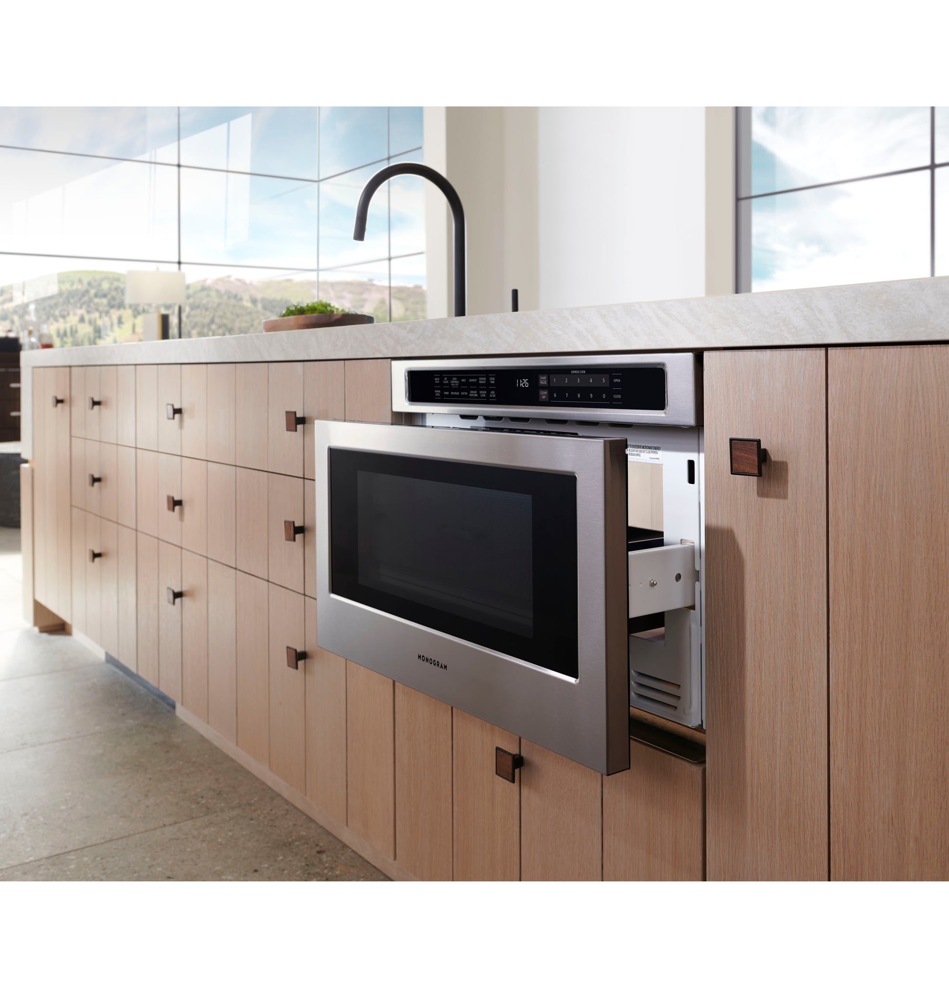 Stainless steel microwave drawer open in light wood built-in kitchen cabinetry with stone countertop and faucet above
