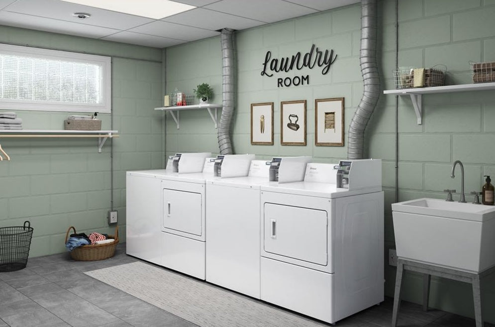 Three white coin-operated electric dryers with rear venting in a spacious laundry room with green walls and utility sink