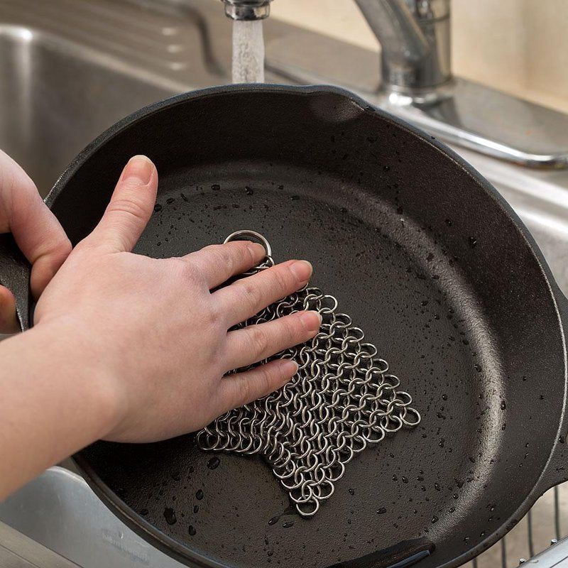 Hand using stainless steel chainmail scrubber to clean cast iron skillet under running kitchen faucet
