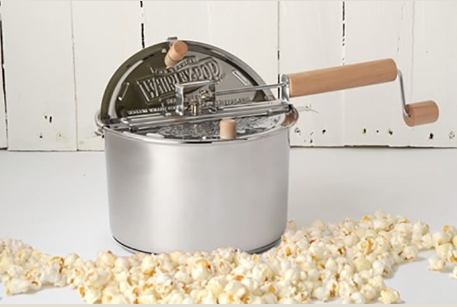 Stainless steel popcorn maker with wooden handled crank and lid, surrounded by popped popcorn on white surface