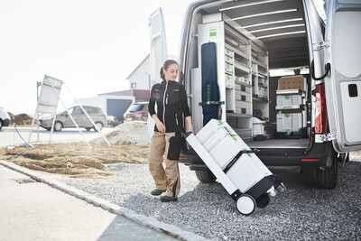 Person transporting white and green Festool storage boxes on a wheeled cart outside a jobsite van for tool organization