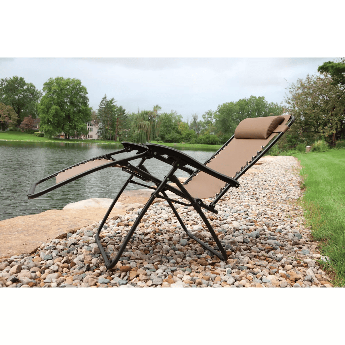 Tan zero gravity convertible lounge chair with padded headrest and black metal frame set on river rocks by a lakeside, ideal for patio furniture and outdoor living spaces.