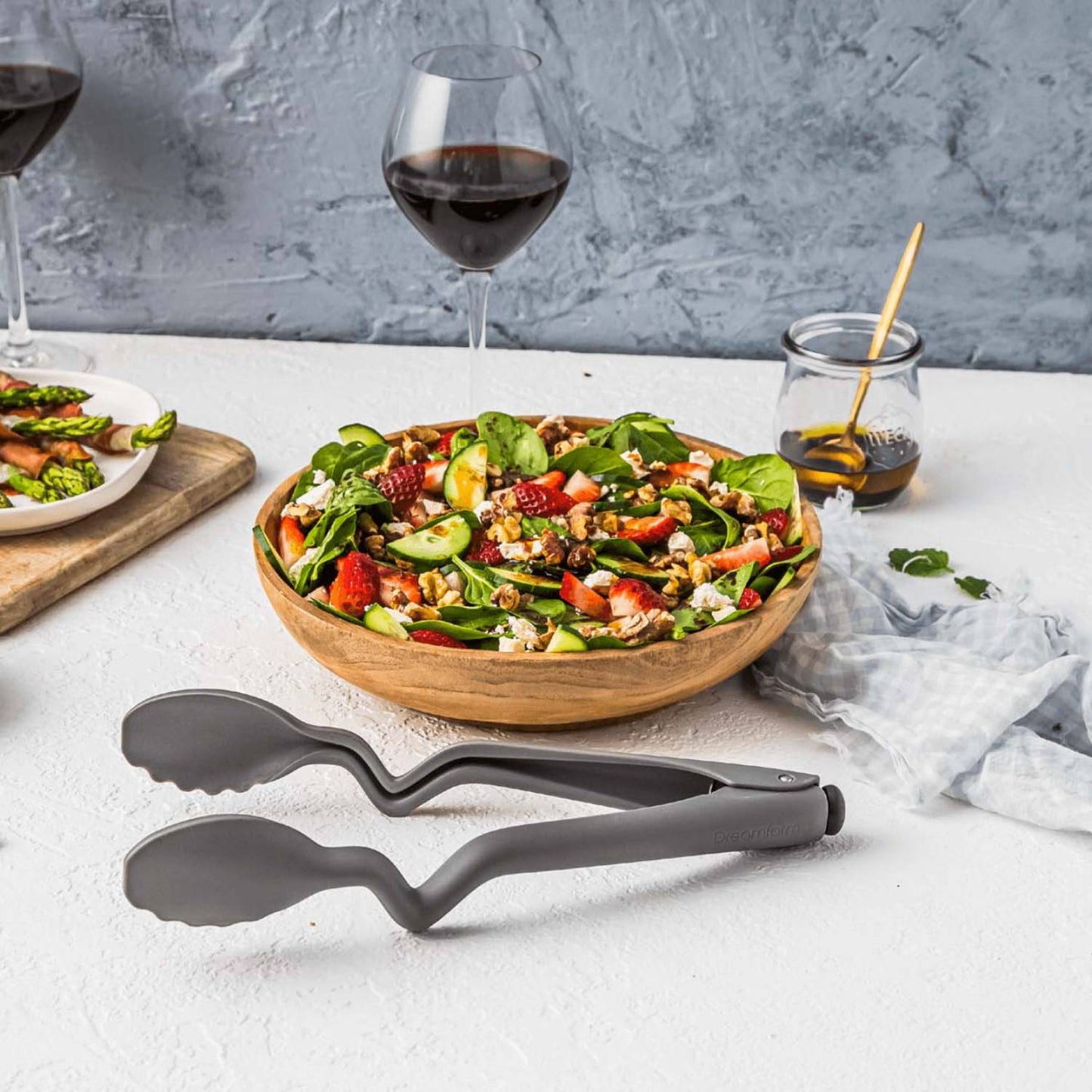 Gray 10.5-inch Dreamfarm kitchen tongs resting beside a wooden bowl of fresh mixed salad on a white surface