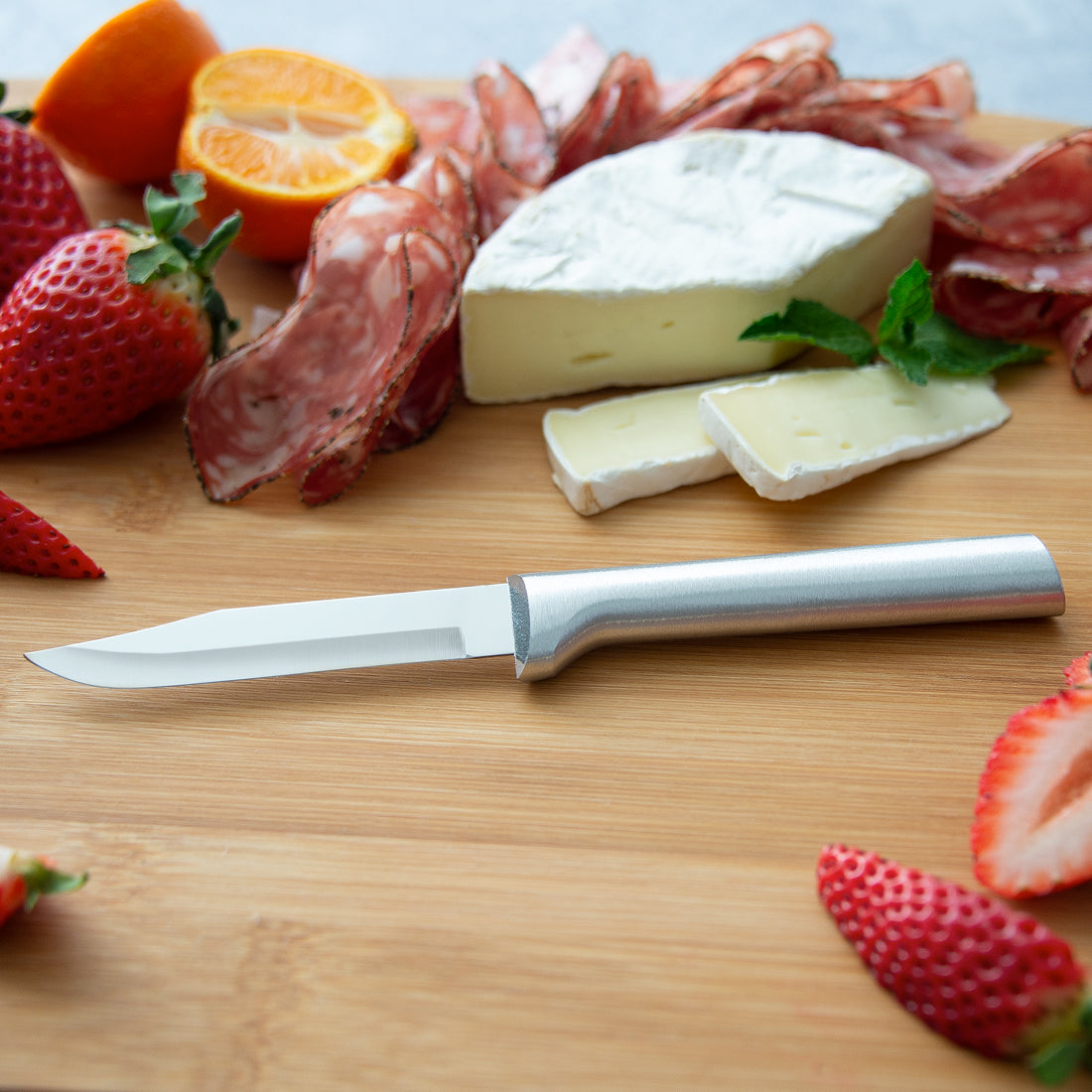 Sharp paring knife with satin finish metal handle on wooden board with cheese, salami, strawberries, and orange slices in kitchenware collections