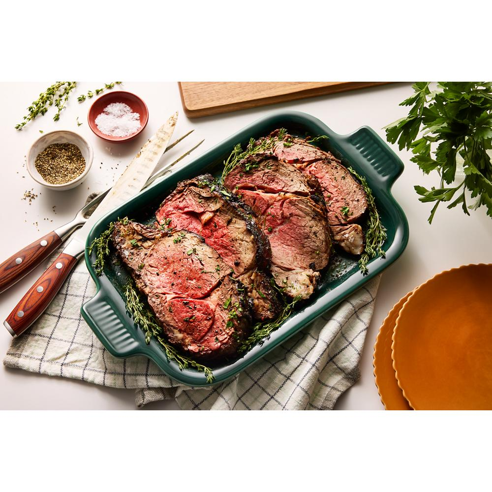Green ceramic baking dish with sliced cooked roast topped with herbs, kitchen knives, and seasoning bowls nearby