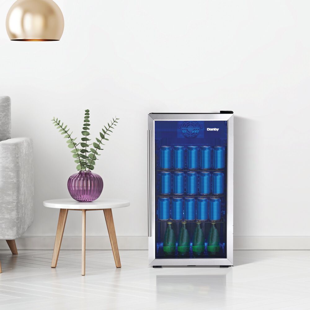 Freestanding beverage center with glass door and wire shelves stocked with assorted cans and bottles in minimalist kitchen setting