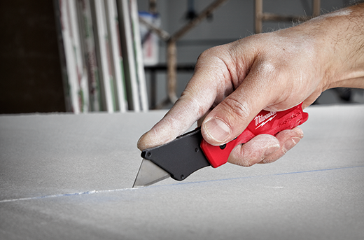 Hand holding red Milwaukee FASTBACK utility knife cutting drywall for woodworking and home improvement tools collection