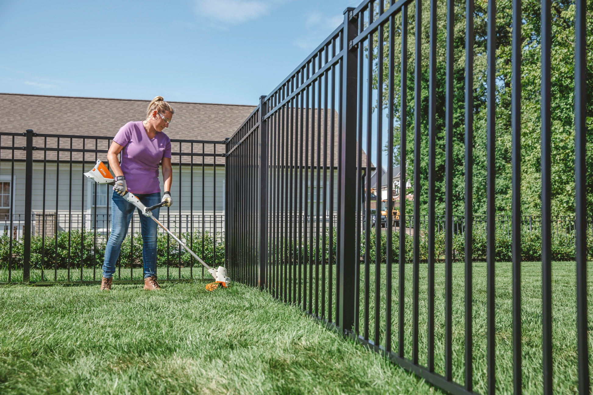 Person using lightweight orange and white battery-powered trimmer along black metal fence for yard care tasks