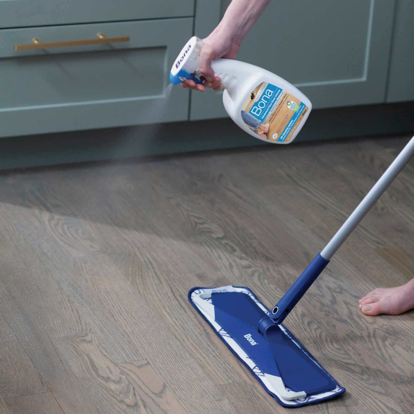 Person applying hardwood floor cleaner spray near blue Bona microfiber mop on wooden floor in kitchen setting