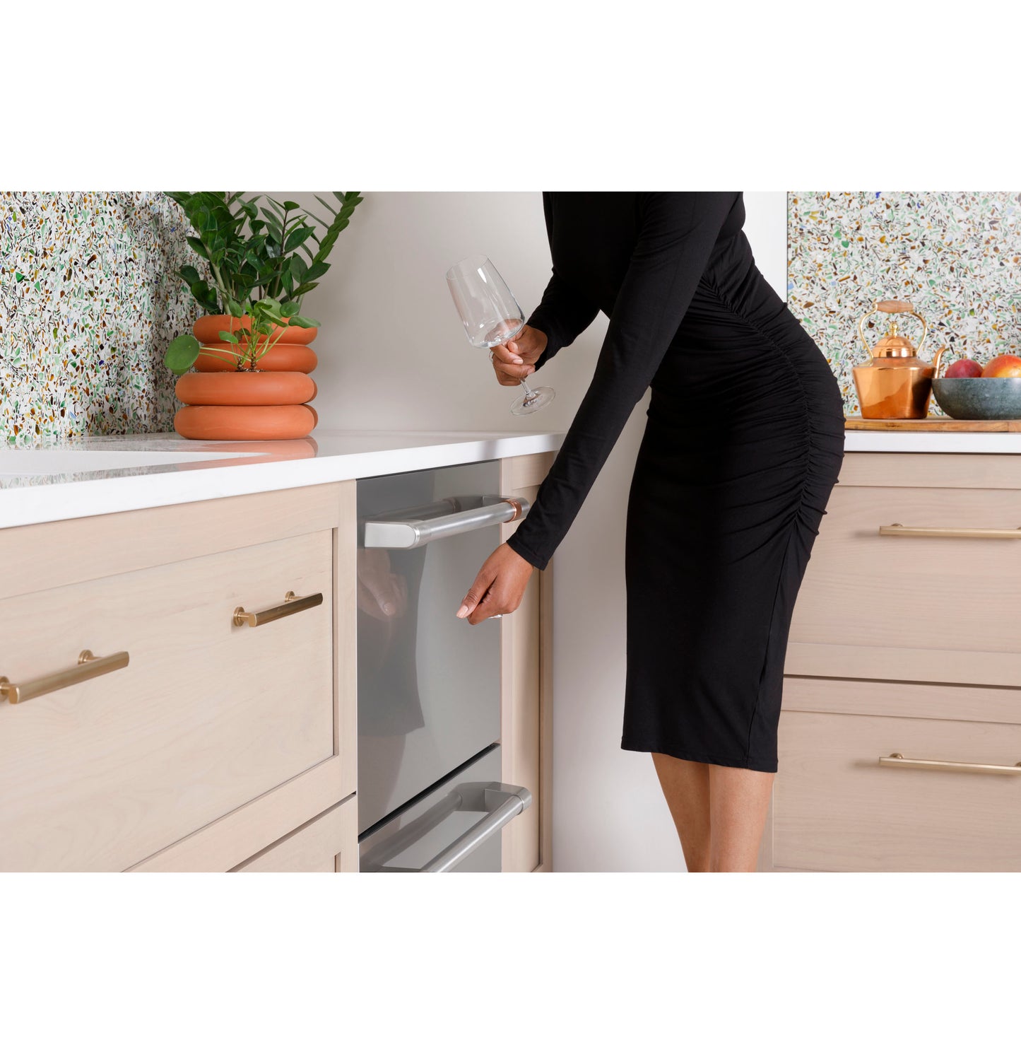 Woman in black dress opening stainless steel dishwasher drawer in modern kitchen with light wood cabinets and terrazzo backsplash