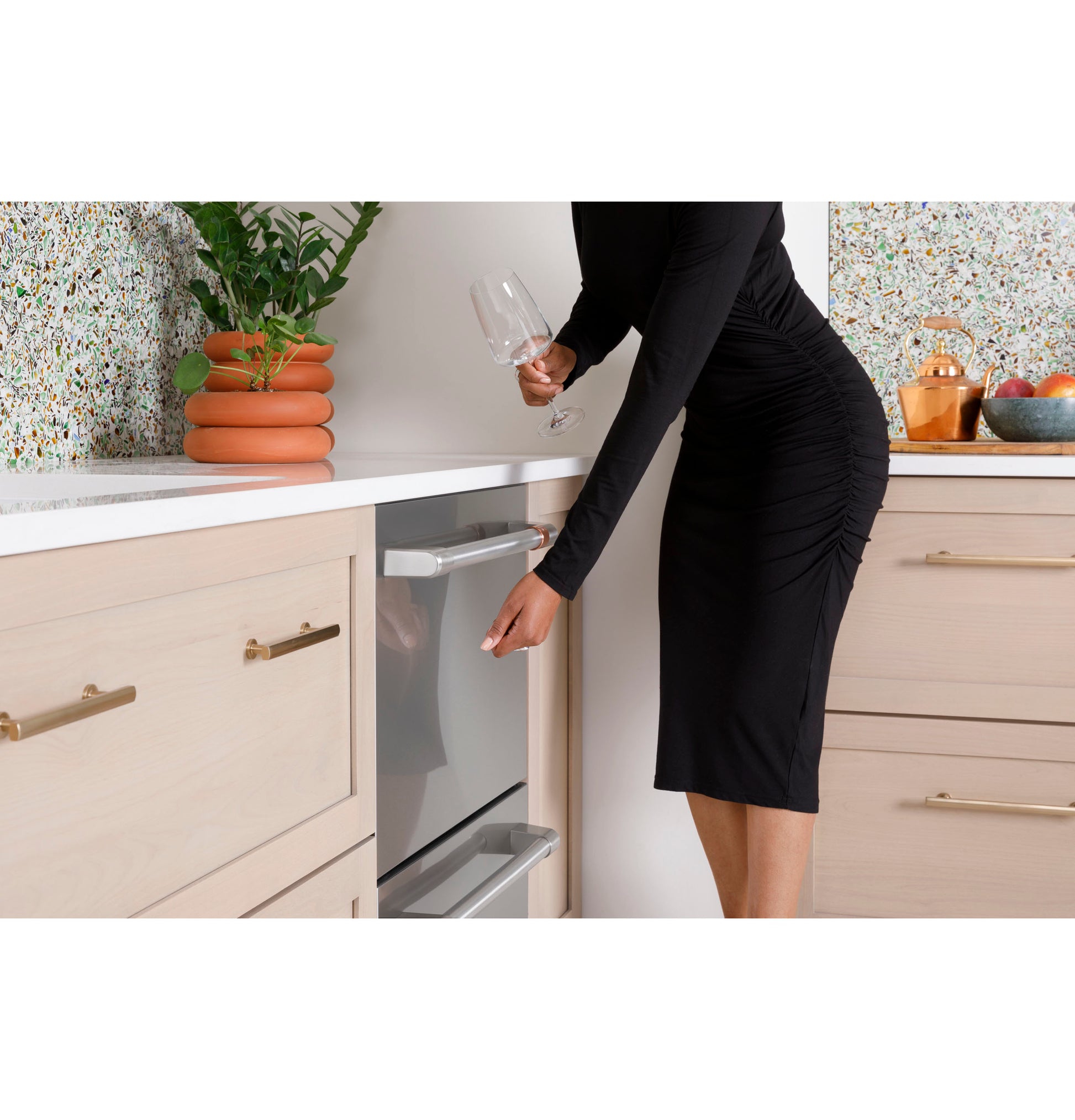 Woman in black dress opening stainless steel dishwasher drawer in modern kitchen with light wood cabinets and terrazzo backsplash
