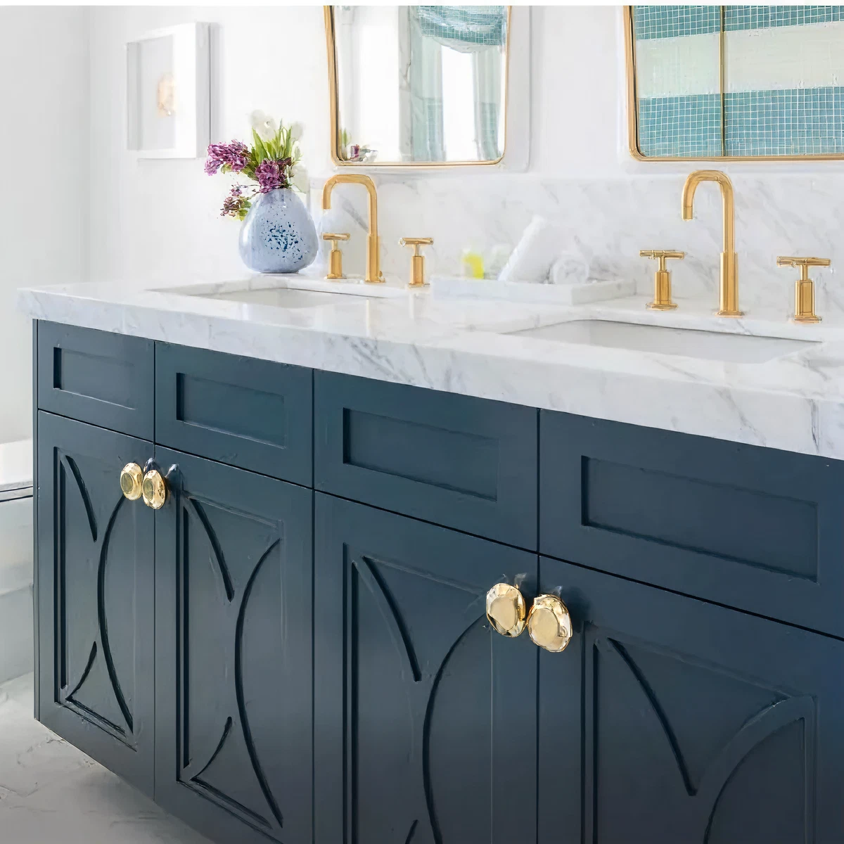 Double-sink bathroom vanity with dark blue cabinetry featuring decorative paneling and large polished brass knobs beneath a white marble countertop with gold-toned faucets and two mirrors.