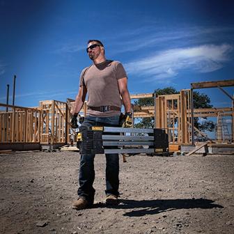Man carrying a compact DEWALT metal folding sawhorse on a construction site with wooden framing in the background