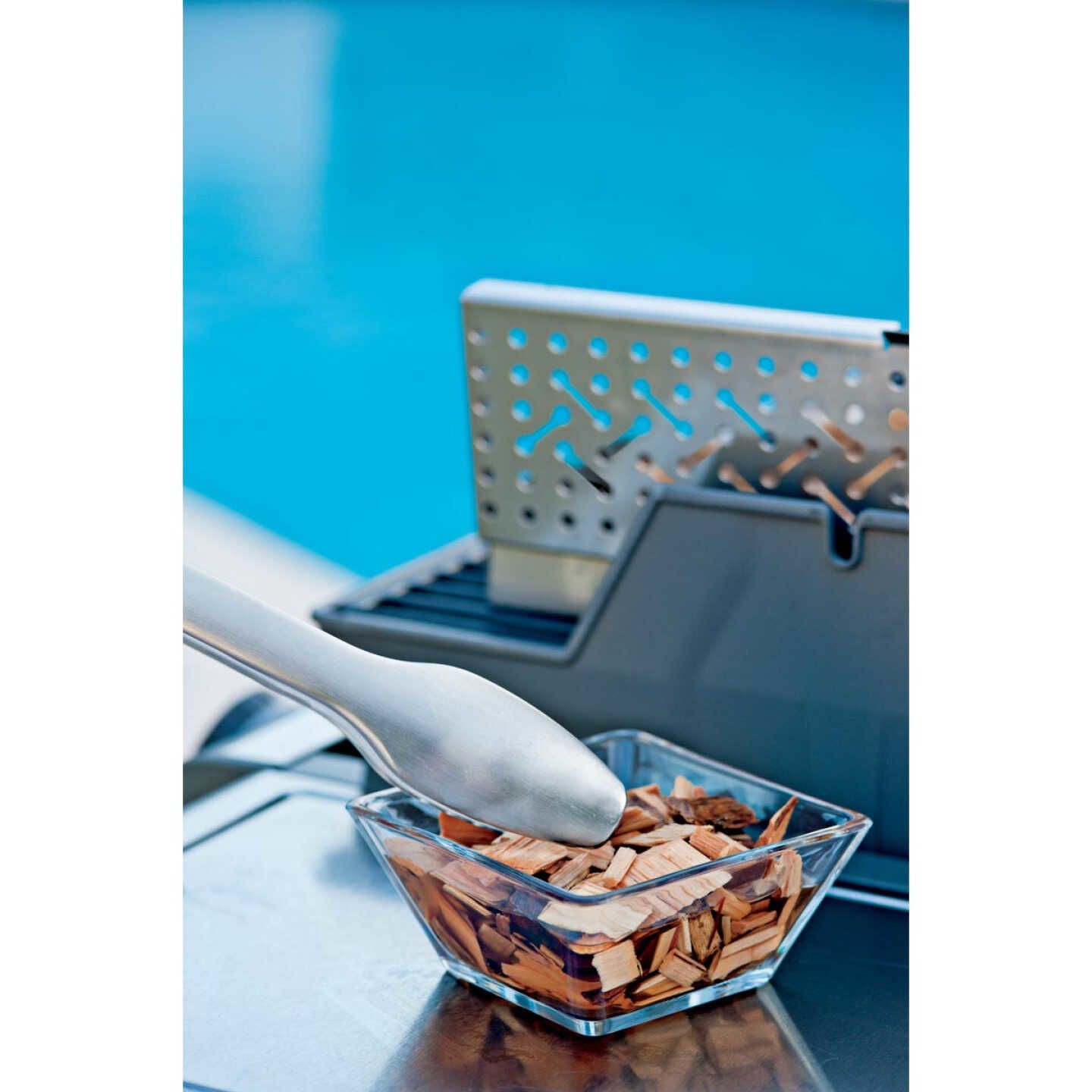 Glass bowl filled with hickory wood smoking chips and stainless steel tongs next to a grill box for outdoor cooking