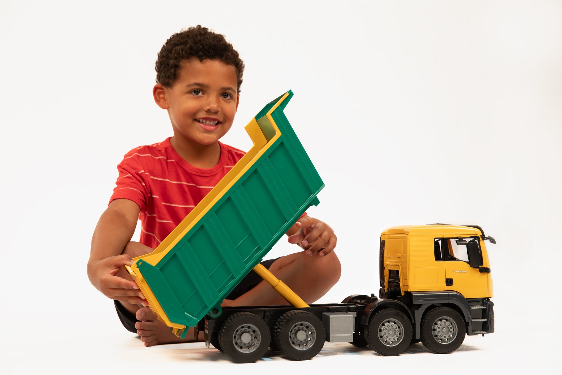 Young boy playing with yellow cab and green bed Bruder Man Dump Truck toy on white background