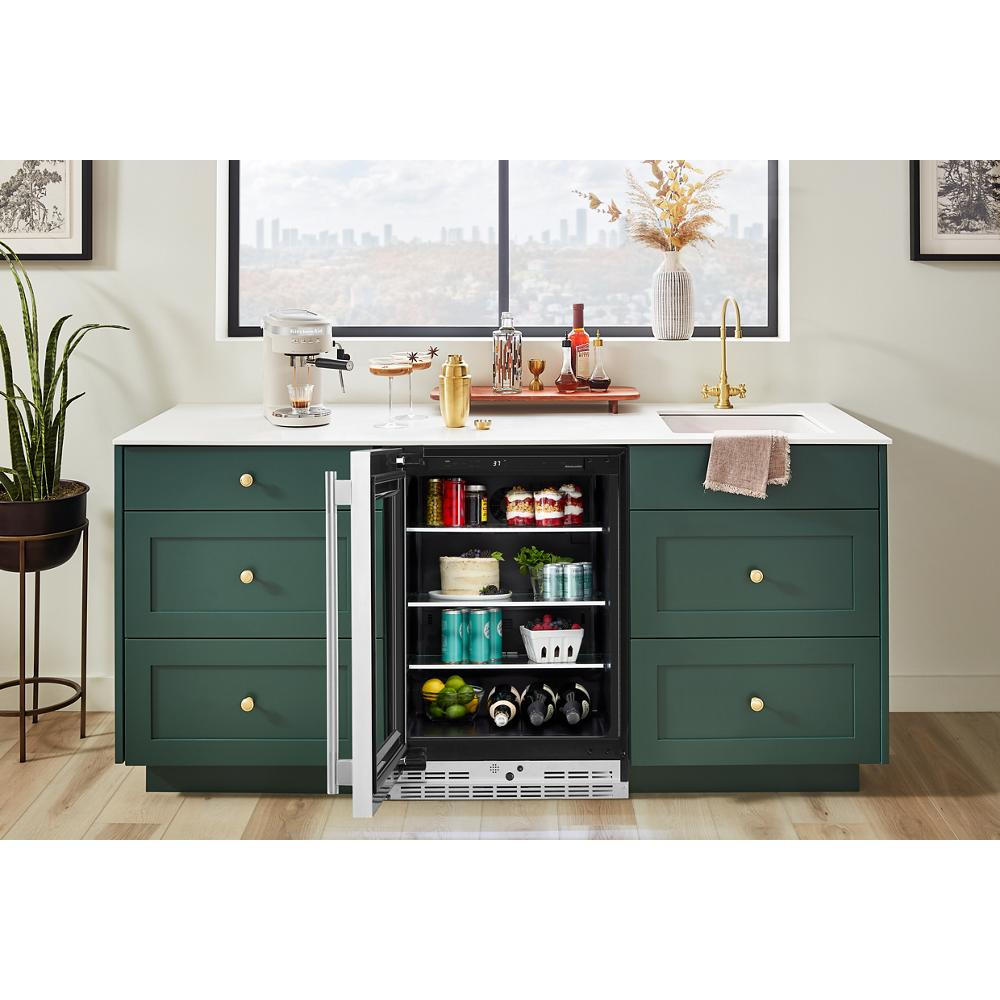 Undercounter refrigerator with glass door showcasing beverages and snacks in modern kitchen with green cabinetry and white countertop