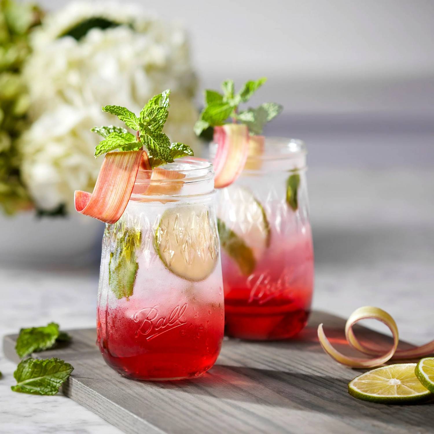 Two clear pint glass jars filled with pink cocktail garnished with mint and rhubarb on kitchenware surface