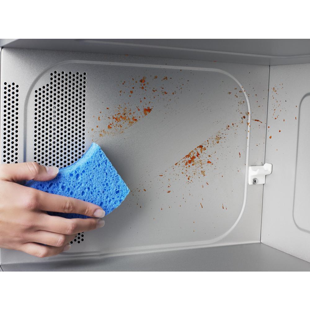 Hand wiping food splatters inside a stainless steel microwave cavity using a blue sponge in kitchen appliances context