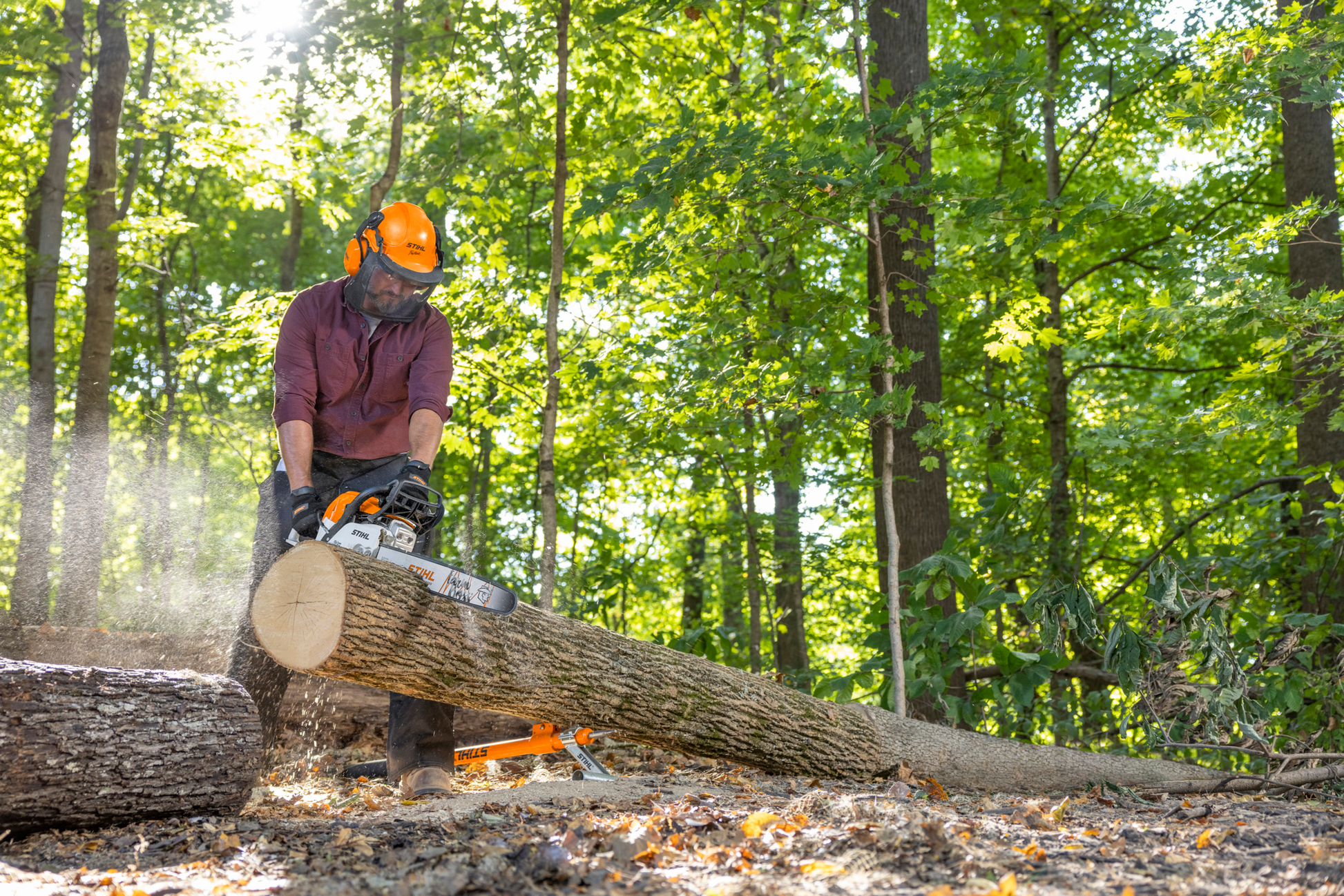 Man wearing safety gear using an orange and white gas-powered chainsaw to cut a large log in a forest setting