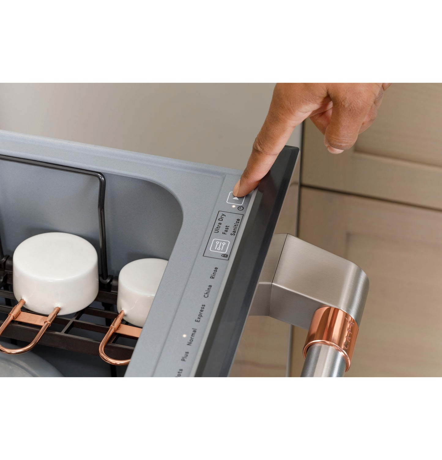 Finger pressing control button on open stainless steel dishwasher drawer with white mugs inside in kitchen