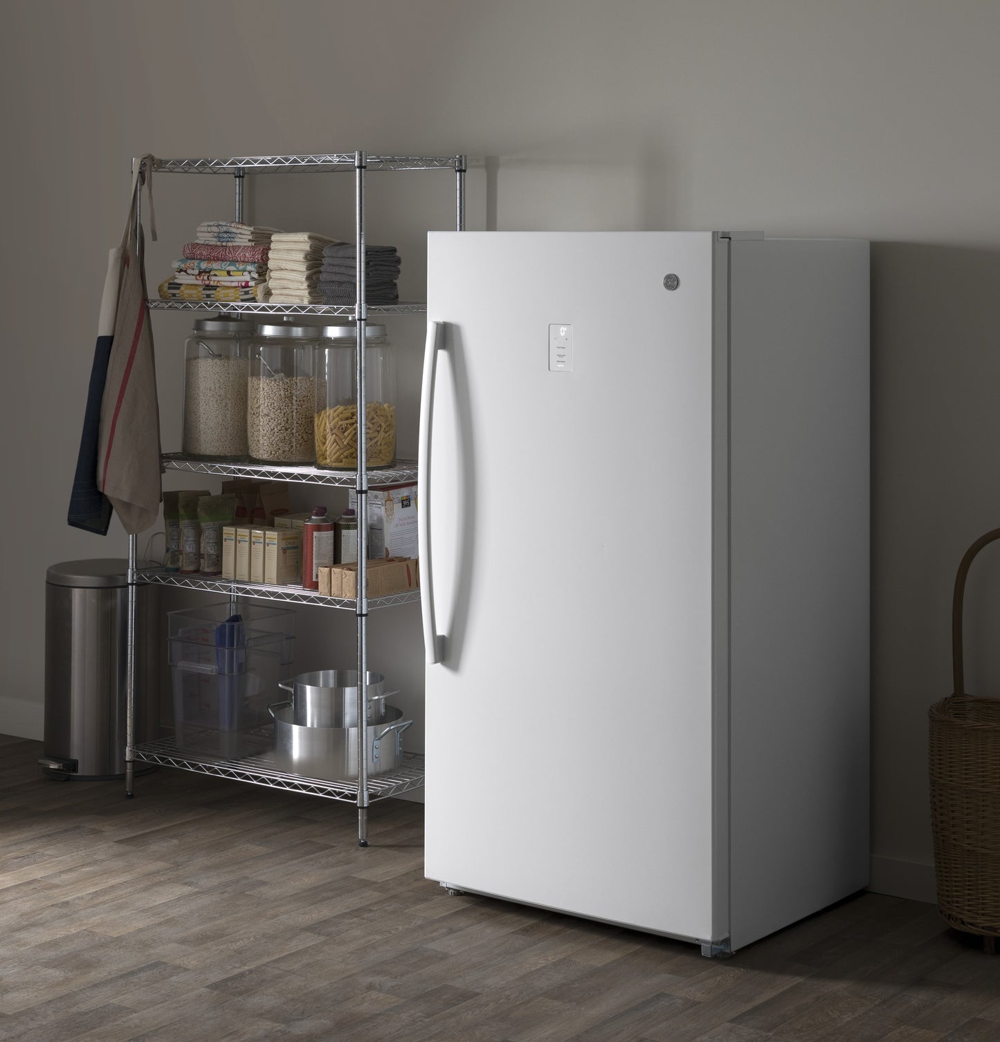White frost-free upright freezer with curved handle in a neutral kitchen setting near wire shelving and storage jars