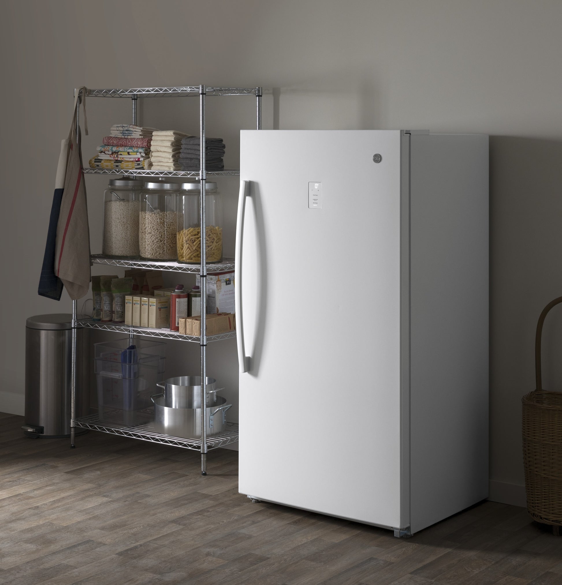 White frost-free upright freezer with curved handle in a neutral kitchen setting near wire shelving and storage jars