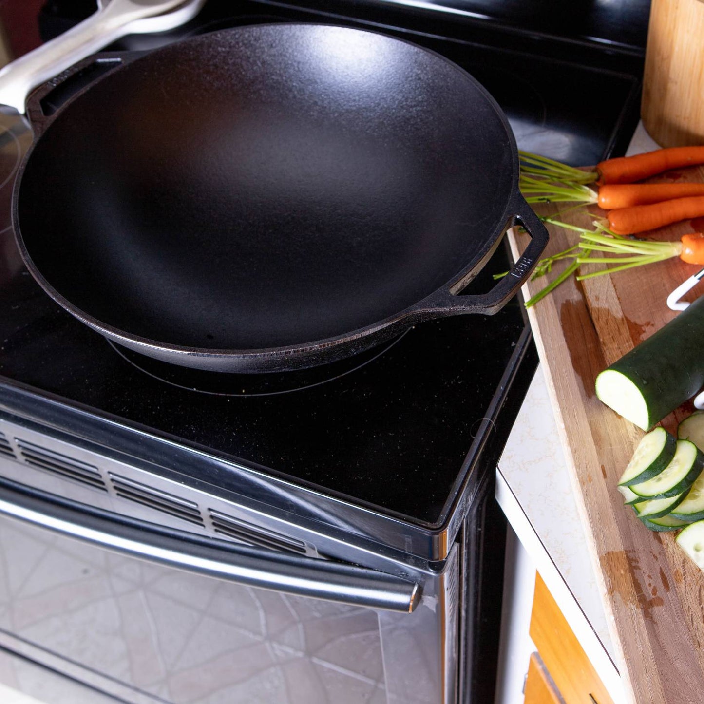Black cast iron wok with wide base and handle on electric stovetop near cutting board with fresh vegetables