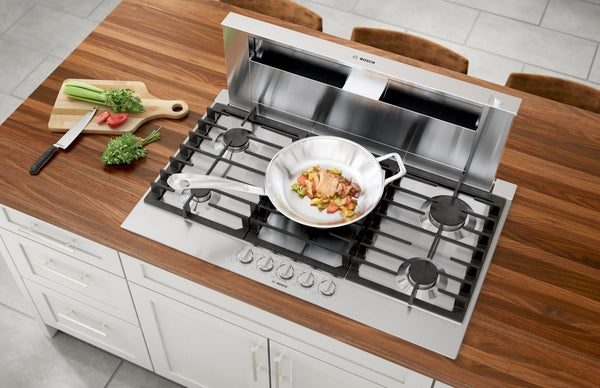 Top view of stainless steel 36-inch gas cooktop with five burners and a pan cooking diced vegetables on wood countertop