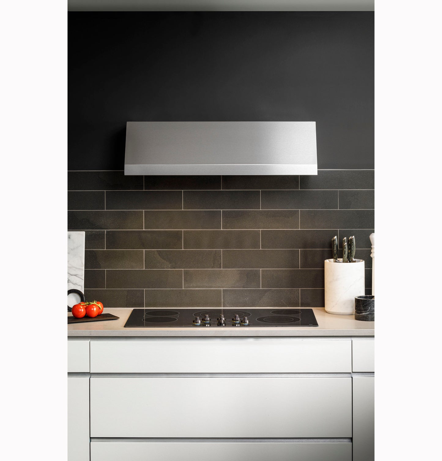 Sleek stainless steel range hood installed above electric cooktop with dark tiled backsplash in modern kitchen setting