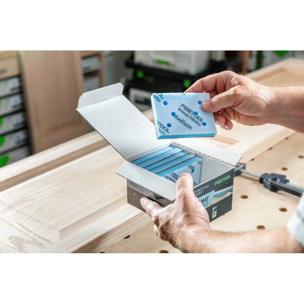 Hands holding a blue foam-backed abrasive pad from an open box of Festool sanding tool accessories on wood surface