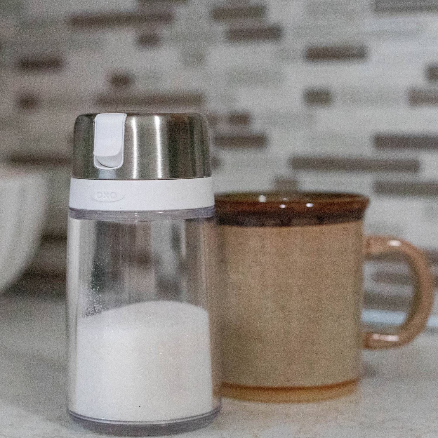 Clear sugar dispenser with stainless steel top and white button on kitchen countertop with ceramic mug nearby