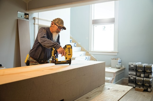 Man using yellow and black cordless narrow crown stapler on wood in bright home improvement setting