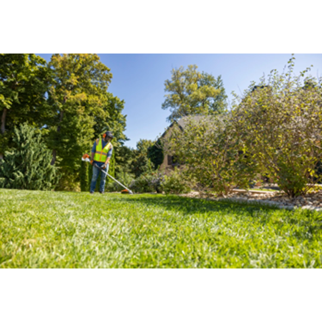 Worker in safety vest using professional trimmer to maintain edges of green lawn near shrubs and residential landscaping