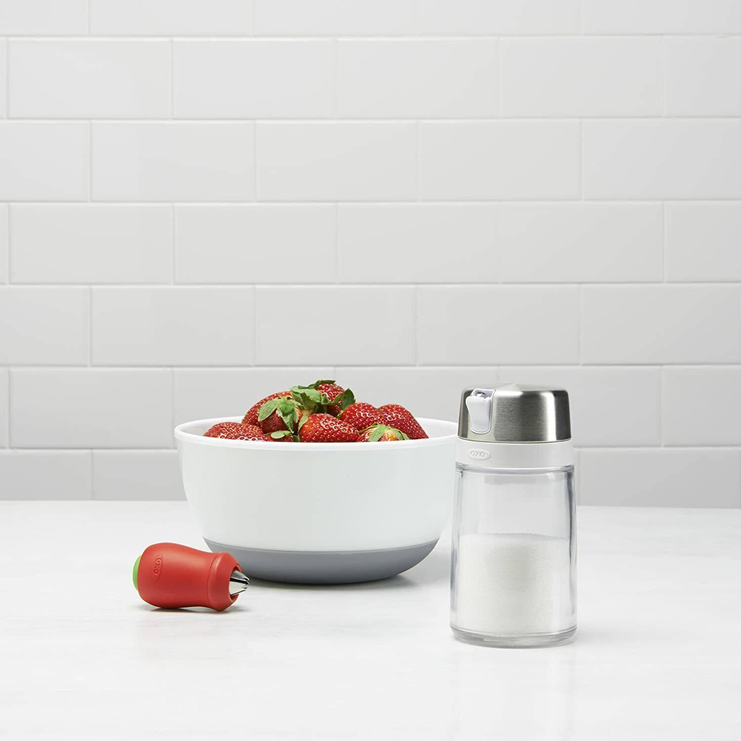 Clear sugar dispenser with stainless steel lid and red adjustable spout next to white bowl of strawberries in kitchenware setting