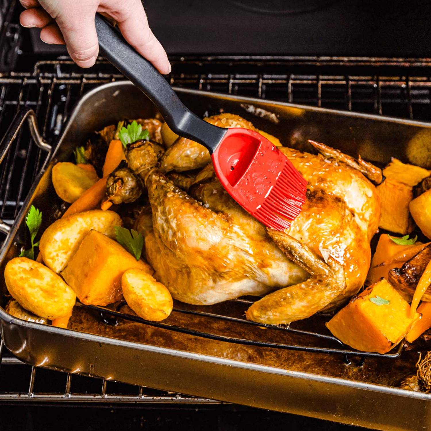 Red silicone basting brush with black handle applying glaze to roasted chicken and vegetables in oven tray from kitchen utensils collection