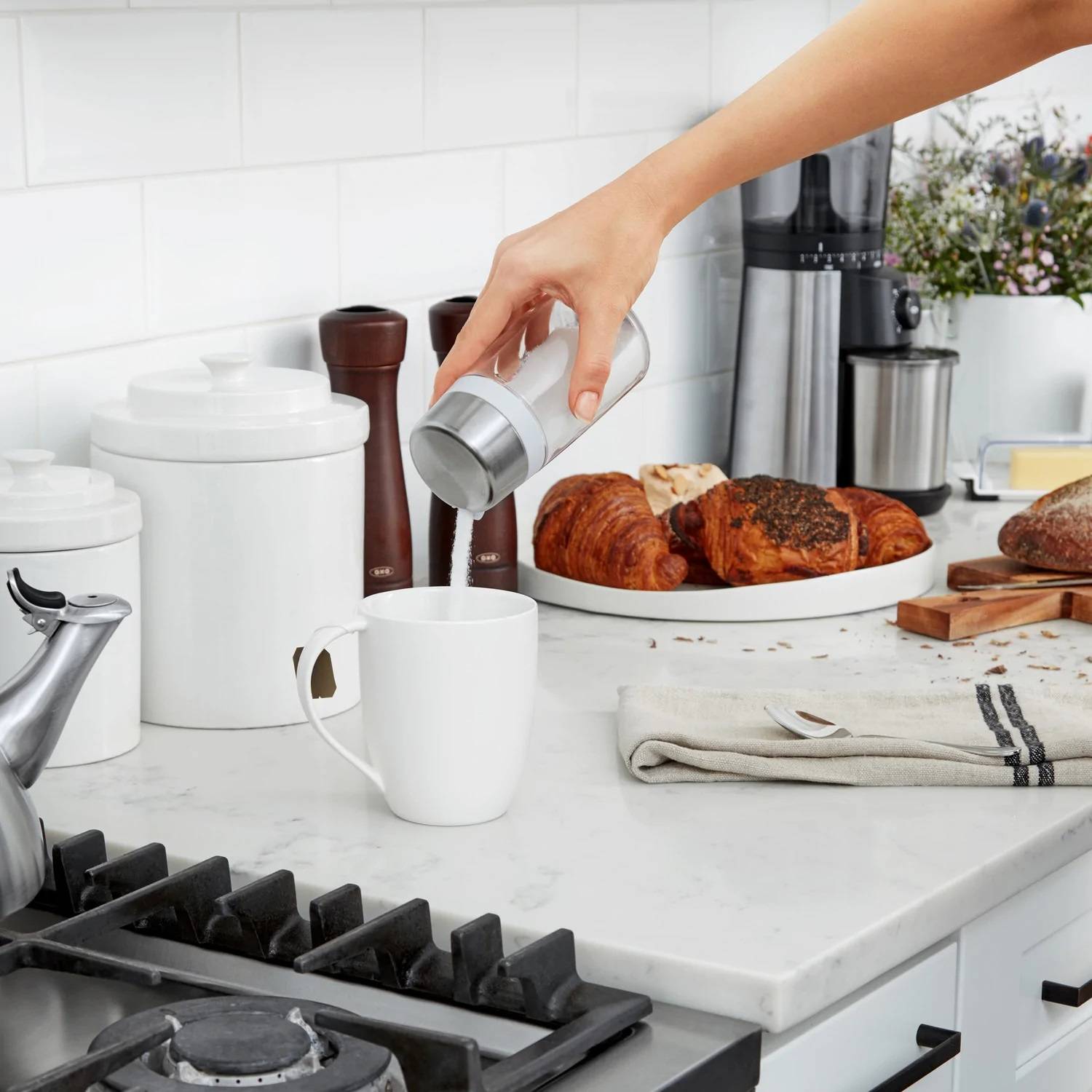 Hand pouring sugar from white and clear dispenser into white mug on kitchen countertop with pastries nearby