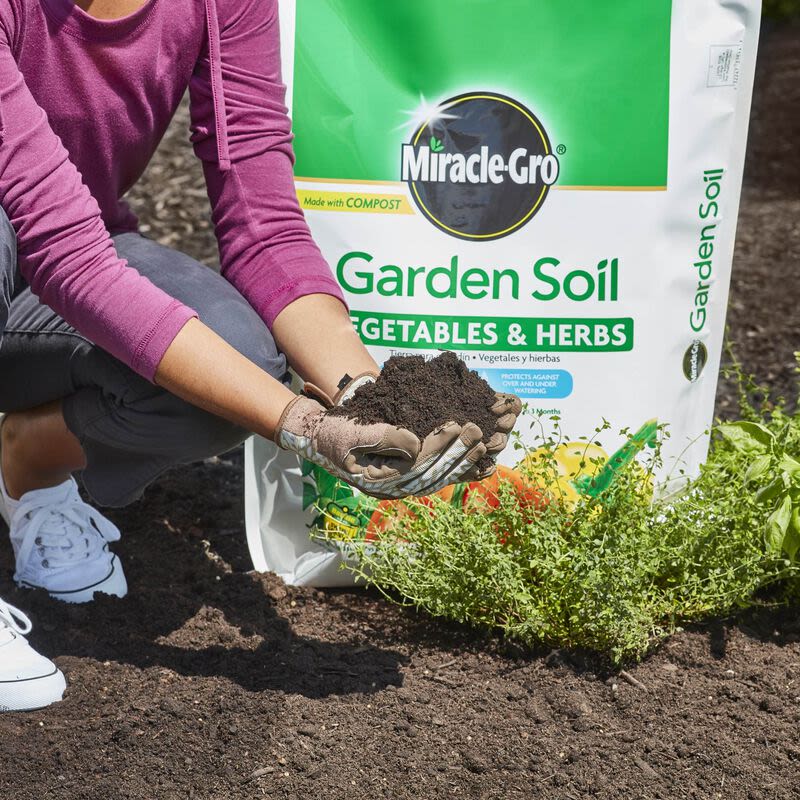 Person wearing gloves holding rich dark soil near a Miracle-Gro garden soil bag in outdoor gardening setting