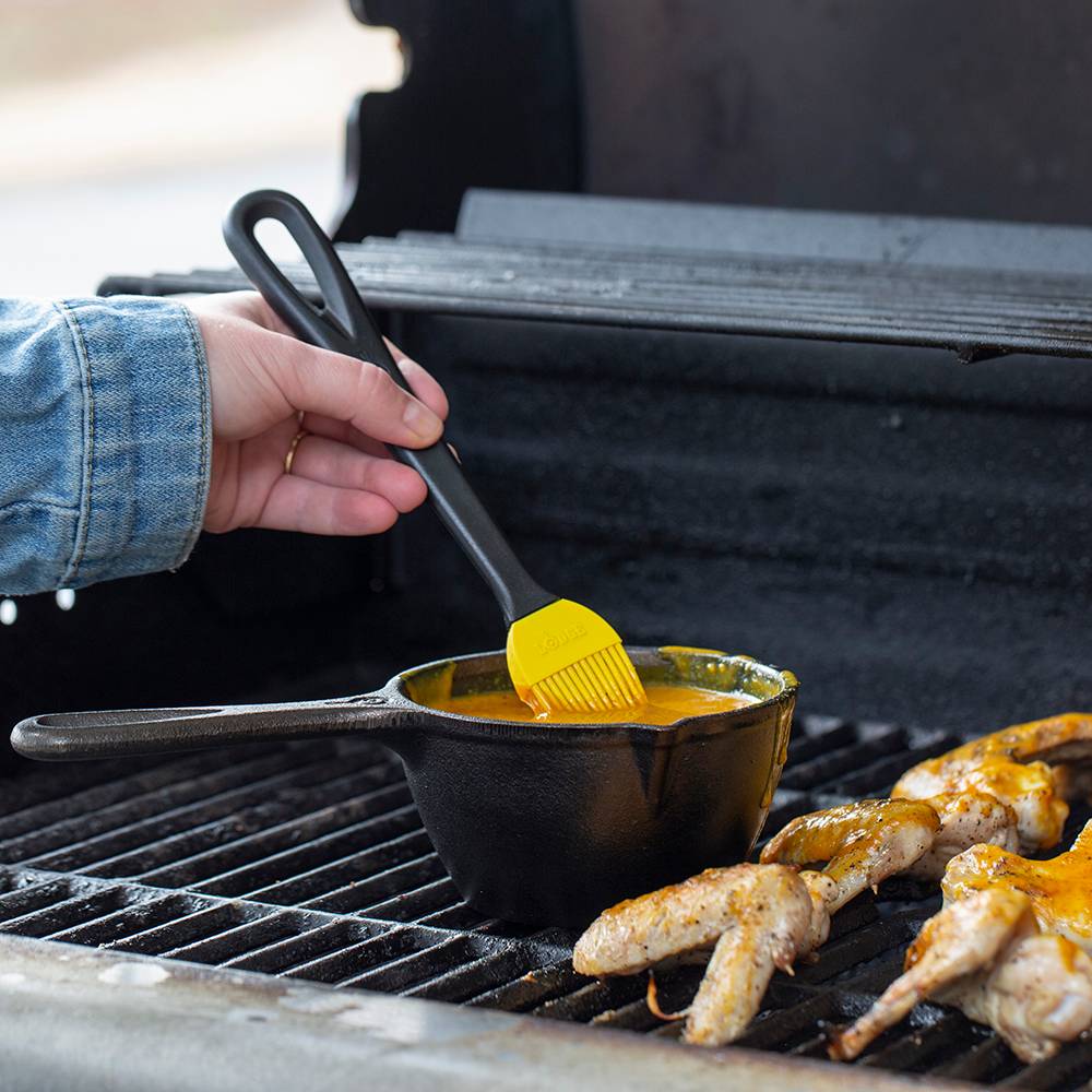 Hand holding yellow silicone brush stirring sauce in black cast iron pot on grill next to cooking chicken wings